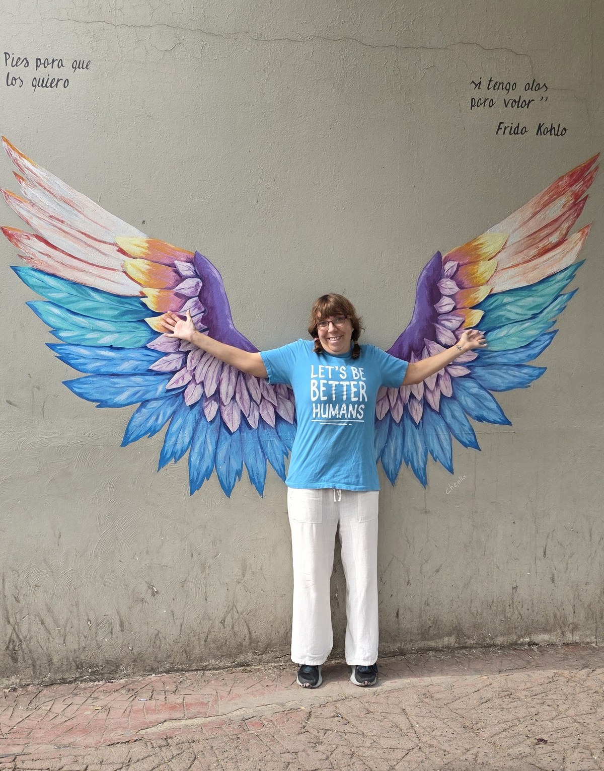 Sherry standing with her arms out in front of a painted mural of rainbow wings, so the wings appear to be her own